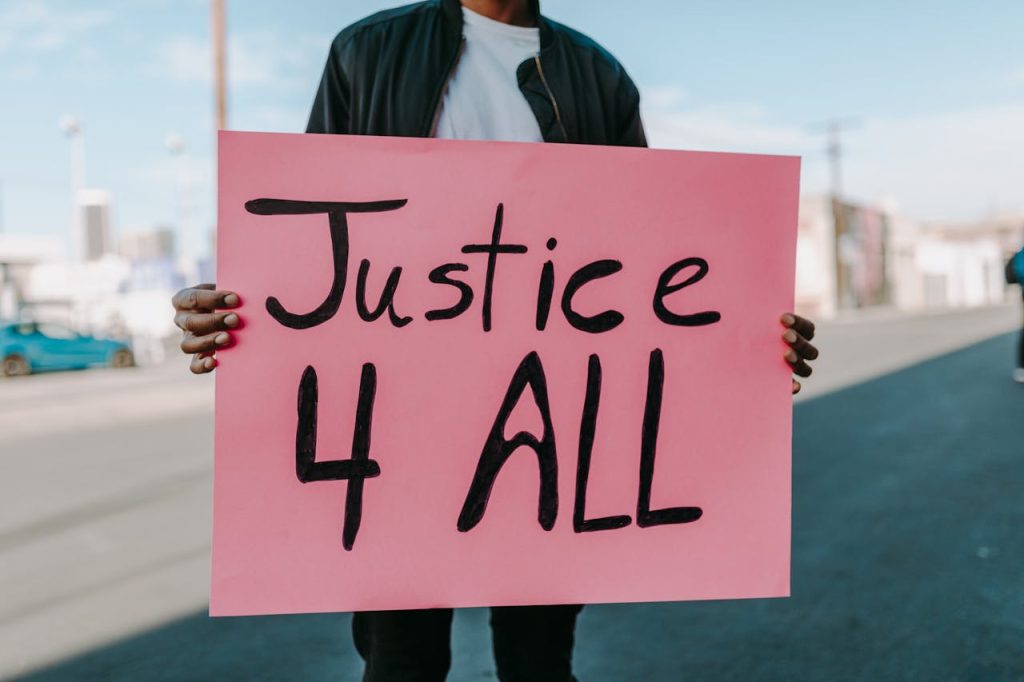 A protester holds a pink 'Justice 4 All' placard on an urban street.